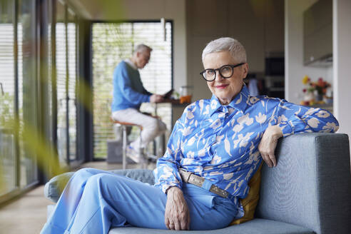 Relaxed senior woman sitting on couch at home with man in background - RBF09231