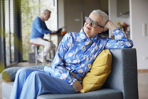 Entspannte ältere Frau sitzt zu Hause auf der Couch mit einem Mann im Hintergrund - RBF09237