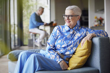 Relaxed senior woman sitting on couch at home with man in background - RBF09238