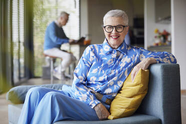 Smiling senior woman sitting on couch at home with man in background - RBF09239