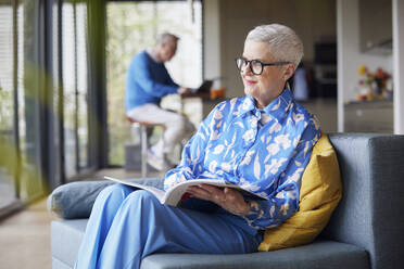 Senior woman sitting on couch at home with magazine - RBF09240