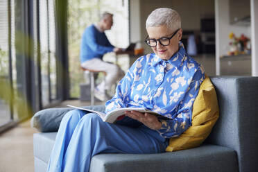 Senior woman sitting on couch at home reading magazine - RBF09241