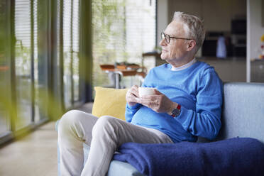 Thoughtful senior man sitting on couch at home with cup of coffee - RBF09243