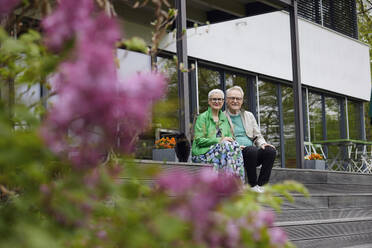 Happy senior couple sitting on steps in front of their home - RBF09256