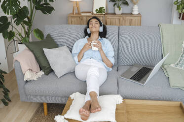 Smiling woman listening to music holding coffee on sofa at home - ALKF00492