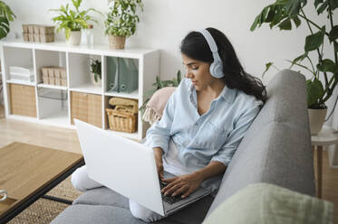 Smiling freelancer wearing headphones using laptop on sofa at home - ALKF00495