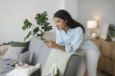 Young woman using smart phone leaning on sofa at home - ALKF00500