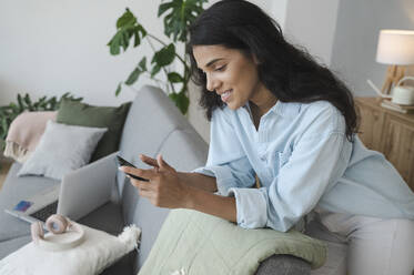 Smiling woman using smart phone leaning on sofa at home - ALKF00501