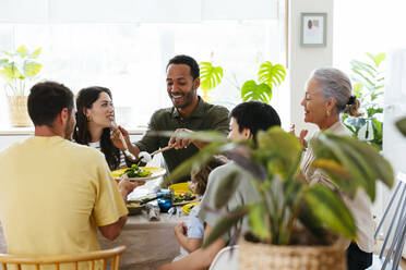 Smiling man serving salad to family at dining table at kitchen - EBSF03692