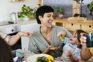 Lächelnder Sohn mit Weintrauben auf dem Schoß der Mutter am Esstisch in der Küche - EBSF03693