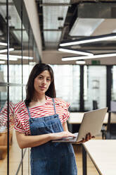 Geschäftsfrau mit Laptop an der Wand lehnend im Büro - PGF01545