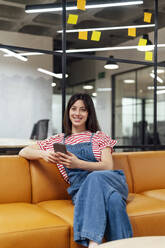 Smiling businesswoman with smart phone sitting on sofa at office - PGF01555