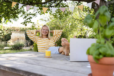 Lächelnder Freiberufler sitzt mit hochgelegten Füßen am Tisch im Garten - SVKF01563
