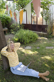 Lächelnder Freiberufler mit Laptop, der sich unter einem Baum im Garten ausruht - SVKF01565
