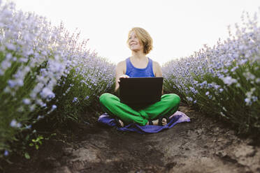 Lächelnde Frau sitzt mit Laptop inmitten eines Lavendelfeldes - SIF00755