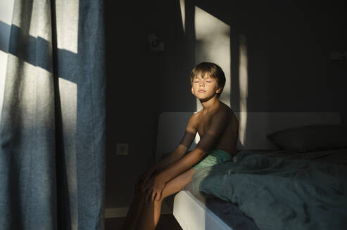 Boy sitting with eyes closed on bed at home - ALKF00504