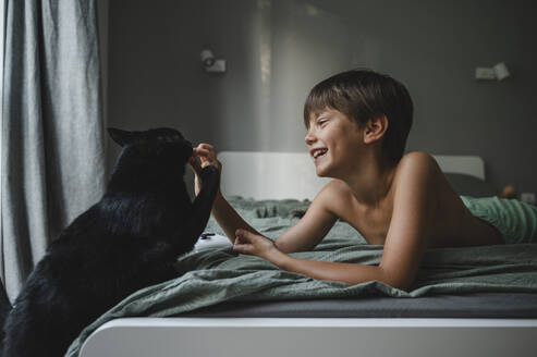 Smiling boy lying on bed and playing with black cat at home - ALKF00508