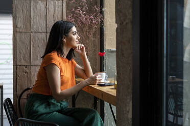Nachdenkliche junge Frau sitzt mit Kaffeetasse und schaut aus dem Fenster eines Cafés - LMCF00463