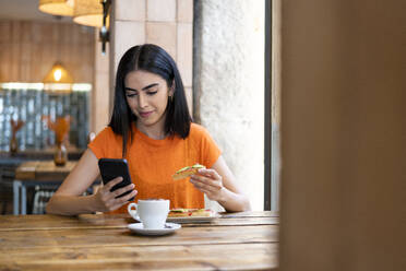 Smiling young woman using smart phone and enjoying food in cafe - LMCF00485