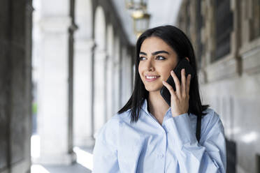 Happy young woman talking on smart phone in colonnade - LMCF00488