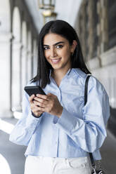Smiling beautiful woman with smart phone standing in colonnade - LMCF00490