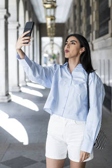 Eine verspielte junge Frau macht ein Selfie, während sie ihre Zunge in einer schönen Kolonnadenumgebung herausstreckt - LMCF00492