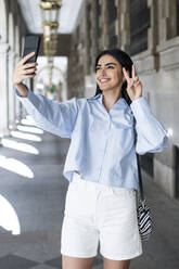 Eine junge Frau macht ein Friedenszeichen und knipst ein Selfie in einer schönen Kolonnade - LMCF00493