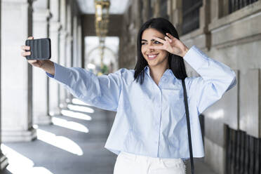Young woman gesturing peace sign and taking selfie in colonnade - LMCF00494