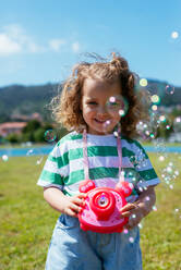 Adorable kleines Mädchen in Freizeitkleidung stehend auf grasbewachsenen Rasen und mit Kamera Bubble Shooter beim Spielen gegen blauen Hintergrund der Natur - ADSF45868