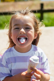 Bezauberndes kleines Mädchen in Freizeitkleidung isst köstliches süßes Eis, während es sich auf einem unscharfen Hintergrund eines Parks amüsiert und in die Kamera schaut - ADSF45869