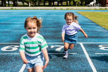 Cute little girls in casual clothes smiling and running on stadium track during outdoor sports training at playground of city park - ADSF45872