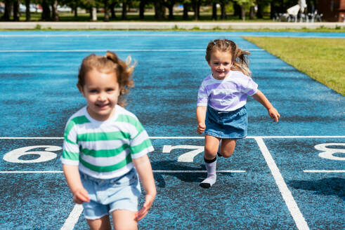 Cute little girls in casual clothes smiling and running on stadium track during outdoor sports training at playground of city park - ADSF45872