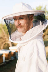 Side view portrait of mature male beekeeper in white protective costume and mask looking at camera while standing in apiary with bees flying around in summer day - ADSF45873