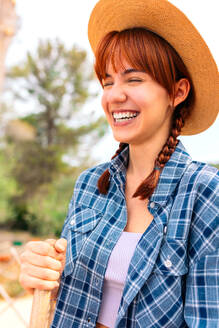 Positive young female in casual checkered clothes with hat smiling and closed eyes while standing with wooden rod near fence in countryside - ADSF45880
