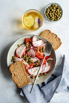 Top view of vegetable salad with organic tomato and capers served in bowl with fork near slices of bread, glass of lemonade and bowl of capers on outdoor table - ADSF45885