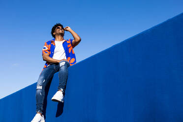 Low angle of young African American male in trendy casual outfit with sunglasses browsing smartphone while sitting looking up on blue concrete fence against blue cloudless sky - ADSF45900