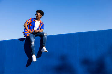 Full body of young African American male in trendy outfit and sneakers with eyeglasses sitting on blue concrete wall against blue sky in daylight - ADSF45901