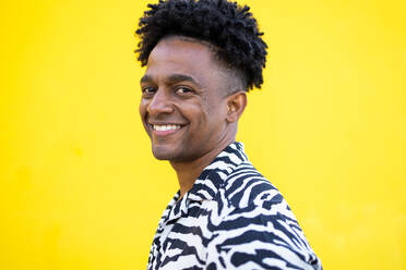 Side view portrait of happy African American male with curly hair hair and trendy casual zebra striped shirt smiling looking at camera while standing on yellow background - ADSF45902