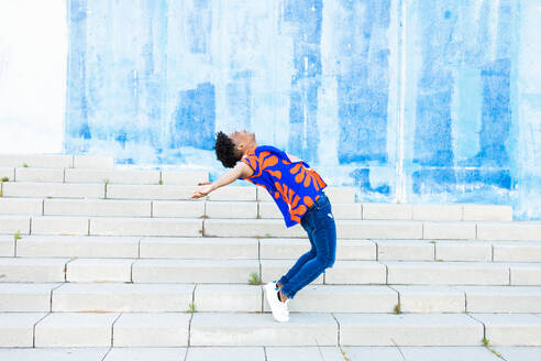 Full body side view of young black male in casual clothes and curly hair jumping above ground in back flip with stretched hands on stairs by blue wall - ADSF45903