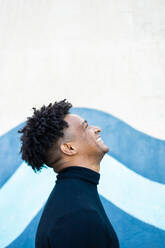 Side view of positive young African American male with curly hair hairstyle in casual clothes standing against blue and white wall while looking up - ADSF45904