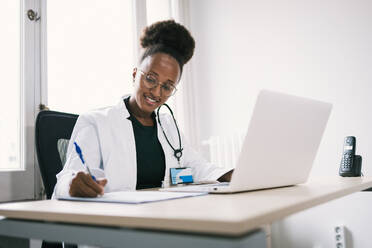 Happy African American female in medical uniform sitting at table with laptop and writing on paper - ADSF45905