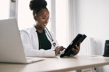 Smiling African American female doctor in eyeglasses, uniform and stethoscope looking at screen of tablet and browsing information with finger while sitting at table with laptop in room in daylight - ADSF45906