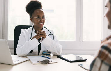 Positive African American female doctor interacting with unrecognizable male patient and looking at each other while sitting at table with laptop, tablet and clipboard in daylight - ADSF45910