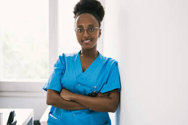 Positive African American female doctor in blue uniform and eyeglasses smiling and looking at camera while standing and leaning on shoulder on wall with crossed hands in room with daylight - ADSF45915