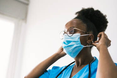 Side view of African American female physician in medical blue uniform with stethoscope looking away while putting on protective mask in hospital - ADSF45917