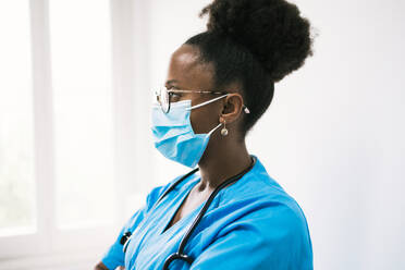 Side view of African American female physician in medical blue uniform and mask with stethoscope looking away thoughtfully in clinic - ADSF45919