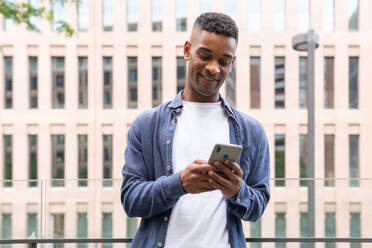 Optimistic African American male entrepreneur in casual outfit smiling and text messaging on mobile phone while working on city street - ADSF45924