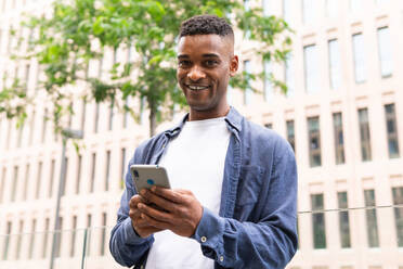 Smiling African American male entrepreneur in casual outfit smiling and text messaging on mobile phone while working on city street and looking at camera - ADSF45925