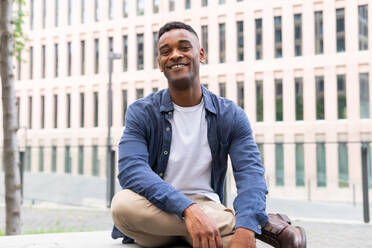 Portrait of friendly African American male in casual clothes smiling and looking at camera while relaxing on bench on city street - ADSF45926