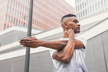 Low angle of serious African American male athlete in sportswear doing warming up exercises during workout and looking away in downtown of city - ADSF45928